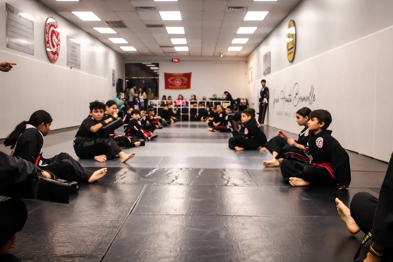 Children in black Galvan’s Martial Arts uniforms sitting along both sides of the mat during a Tae Kwon Do class, with parents watching from the back of the room