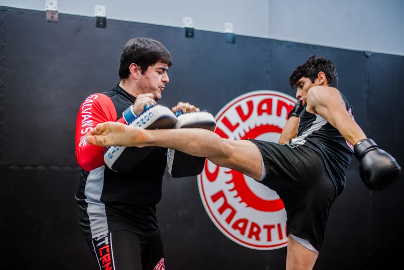 A Galvan’s Martial Arts coach braces a pair of focus pads while a student lands a high kick, in front of the red Galvan’s dragon logo painted on the gym wall